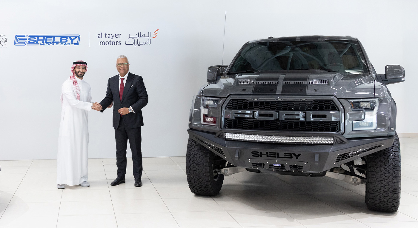 Two men in formal attire shake hands beside a grey Shelby truck in a showroom. Logos for Shelby and Al Tayer Motors are visible on the wall behind them.